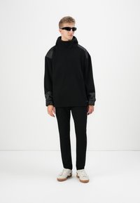 Felpa nera oversize con spalle testurizzate, abbinata a pantaloni neri slim-fit. Accessori comprendono occhiali da sole neri e sneakers beige.