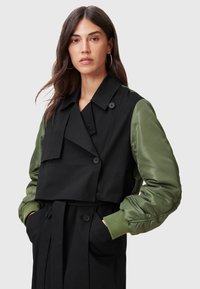 Sort trenchcoat med grønne bomberærmer, der har et dobbeltbrystet design, store knapper og et bælte i taljen. Glat overflade.