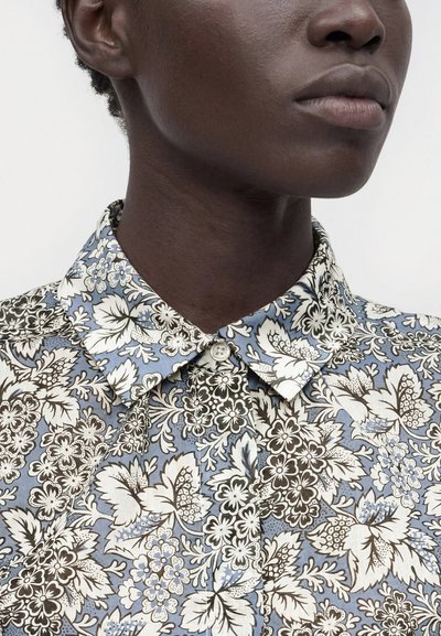 Personne à la peau foncée portant une chemise boutonnée avec un motif floral bleu, blanc et noir, visible du cou au menton et à l'oreille.