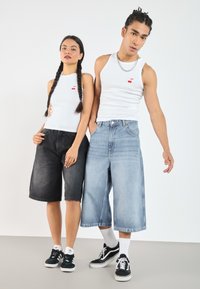 Dva modela nosita usklajene bele majice s češnjevim vezenjem. Eden nosi črne jeans kratke hlače, drugi pa svetlo modre hlače do kolen.