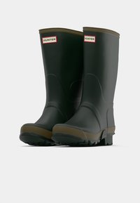 Μαύρες καουτσούκ γαλότσες με καφέ σόλα, φέρουσες μια υφασμάτινη κορυφή με υφή και το λογότυπο "HUNTER" σε μια κόκκινη και λευκή ετικέτα.