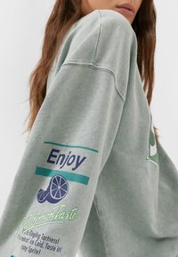 Miętowozielony, oversize'owy sweter z ribbami przy mankietach. Posiada kolorowe grafiki na rękawie, w tym tekst oraz wzór plasterka cytryny.