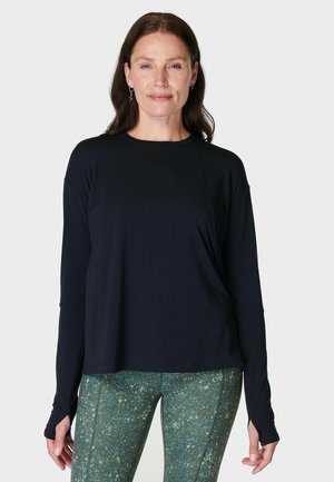 Vrouw met lang bruin haar draagt een zwart shirt met lange mouwen en groene legging met vlekken, staand tegen een effen lichtgrijze achtergrond.