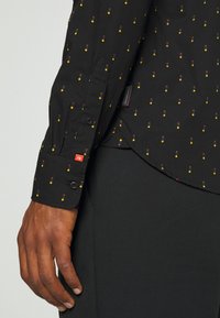 Camisa negra de botones con un pequeño patrón colorido de puntos rojos, amarillos y verdes. La tela es liviana y tiene una textura suave.