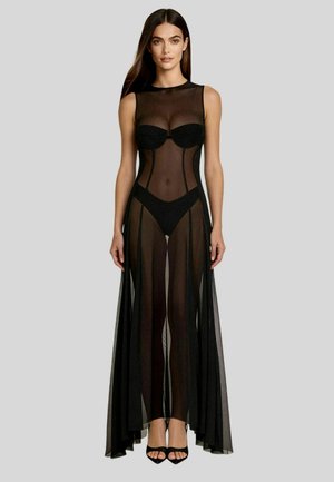 Donna che indossa un abito nero trasparente senza maniche con biancheria intima nera aderente e tacchi neri aperti, in piedi davanti a uno sfondo grigio.