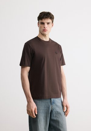 Ung mann står vendt mot kamera iført en ensfarget, mørkebrun t-skjorte med korte ermer og lyseblå denimjeans mot en ensfarget bakgrunn.