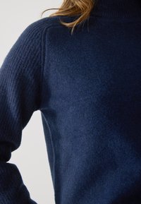 Pulover cu guler înalt de culoare albastru marin, cu textură încrețită pe umeri și mâneci; material moale, gros, cu o finisare netedă.