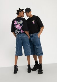 Camisetas negras de gran tamaño con estampados gráficos, combinadas con pantalones cortos de carga de denim azul que tienen bolsillos laterales y un ajuste relajado. Calzado negro.