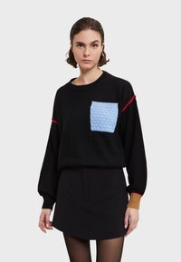 Maglione nero con una tasca testurizzata blu; presenta un accento rosso su una manica e dettagli a costine a contrasto nei polsini. Indossato con una gonna nera.