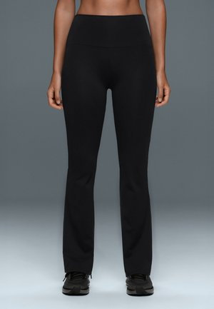 Personne portant un pantalon noir cintré taille haute et des baskets noires, debout devant un fond gris, le torse et les jambes visibles.