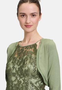 Zielona sukienka z misternym haftem kwiatowym i cekinami na przezroczystym materiale, w połączeniu z pasującą miękką zieloną bolerkiem.