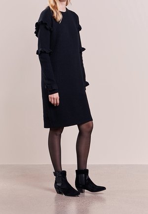Frau trägt ein langes schwarzes, geripptes Kleid mit Rüschenärmeln, schwarze transparente Strumpfhosen und schwarze Stiefeletten mit Schnallen.