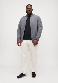 Chaqueta bomber gris con cierre frontal, puños acanalados y un bolsillo lateral con cremallera, llevada sobre una camisa negra, acompañada de pantalones blancos y zapatillas negras.