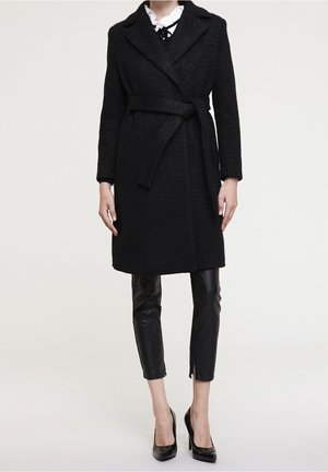 Femme portant un manteau noir texturé ceinturé sur un chemisier blanc et un pantalon en cuir noir raccourci avec des talons noirs, sur fond uni.