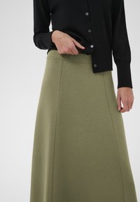 Falda verde oliva en línea A hecha de tela suave, con una textura lisa y un detalle de costura. Combinada con un cárdigan negro.