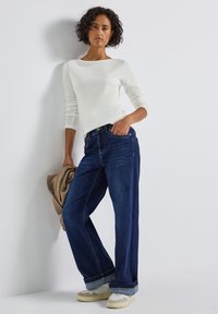 Un haut à manches longues blanc associé à un jean ample bleu foncé, avec un ourlet roulé et des baskets de couleur claire, avec un vêtement beige drapé sur le bras.