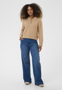 Bēša adīta džemperis ar apkakles izgriezumu un trim pogām, apvienots ar platām zila denim džinsām un gaišām kurpēm.