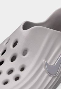 Chaussure à enfiler gris clair avec des trous de ventilation et un logo Nike, présentant une surface texturée et un bord supérieur contouré.