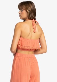 Koralfarvet cropped top med volanger og hæklet detaljer. Binder bagpå med frynser. Sættes sammen med matchende løstsiddende bukser.