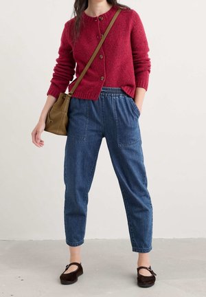 Femme portant un pull rouge boutonné, un pantalon en jean bleu taille haute avec de grandes poches avant, des chaussures Mary Jane marron et un sac à bandoulière marron.