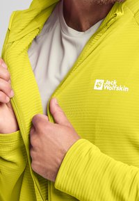 Giacca zip-up verde lime con tessuto testurizzato, dotata di colletto alto e con il logo "Jack Wolfskin" in bianco sul petto.