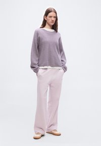 Γυναίκα που φοράει πουλόβερ από λεπτή πλέξη σε λιλά χρώμα, ροζ ανοιχτό παντελόνι με φαρδιά πόδια και μπεζ αθλητικά παπούτσια, στέκεται μπροστά σε απλό λευκό φόντο.