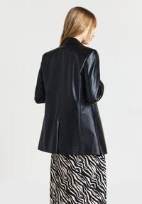 Donna con capelli biondi che indossa un blazer in pelle nera e una gonna a strisce zebrate in bianco e nero, vista di spalle su uno sfondo neutro.