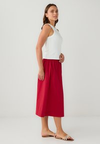 Haut sans manches blanc associé à une jupe midi rouge. La jupe présente une taille froncée et une texture lisse, portée avec des sandales légères.