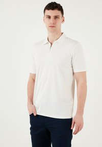 Weißes Polo-Shirt mit kurzen Ärmeln, Reißverschluss und klassischem Kragen. Sanfte, glatte Textur und entspannte Passform. Getragen mit dunklen Hosen.