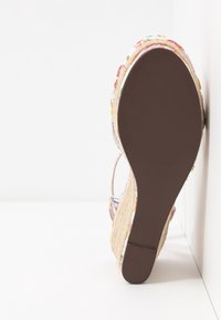 Sandale compensée fleurie avec semelle marron, talon enveloppé de jute et bride à la cheville, positionnée droite contre un mur et un sol blancs.