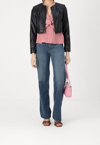 Donna che indossa una giacca di pelle nera corta, un top rosa annodato, jeans blu, sandali marroni con borchie e tiene in mano una piccola borsa rosa.