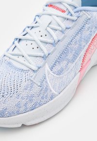 Gros plan d'une basket tricotée bleu clair et blanche avec lacets blancs, détail de couture rouge et semelle blanche mouchetée sur fond blanc.