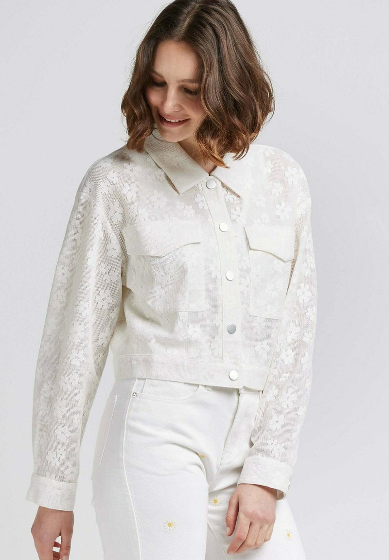Veste en denim blanche cropped avec un texture de dentelle florale, deux poches poitrine et des boutons en argent, associée à un jean blanc orné de marguerites.