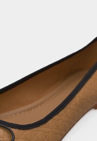 Gros plan sur une chaussure plate couleur sable avec des motifs floraux embossés et un bord noir le long de l'ouverture.