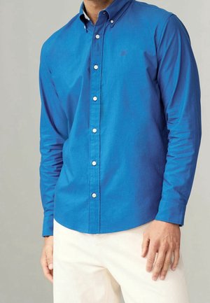 Homme portant une chemise bleue ajustée à manches longues avec boutons et un pantalon crème, debout avec une main détendue le long du corps.