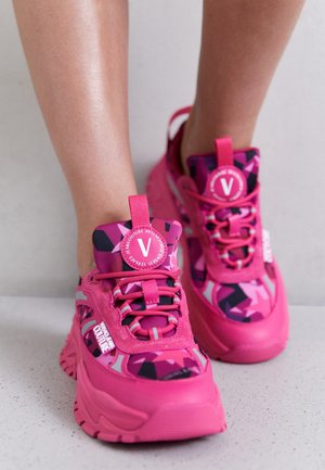 Ben iført lyserøde Versace Jeans Couture sneakers med geometriske sorte og pink mønstre, snørebånd bundet, stående på plettet gulv.