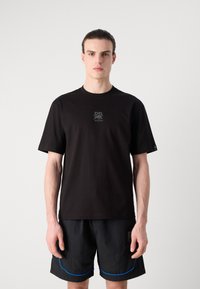T-shirt nera in cotone con maniche corte e scollatura rotonda, con un piccolo logo grigio sul petto. Indossata con pantaloni corti neri.