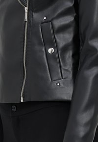 Veste en cuir noir avec une fermeture éclair argentée, un accent de bouton-pression et une pochette sur la poitrine. Caractérisée par une texture lisse et un design ajusté.