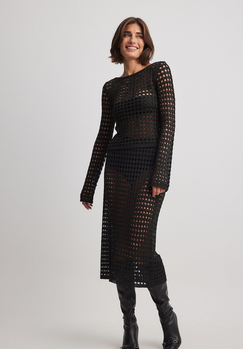 Schwarzes gehäkeltes Kleid mit langen Ärmeln und knielangem Saum. Das Design zeigt offene Rautenmuster und eine figurbetonte Silhouette.