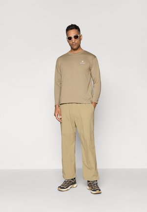 Beige Langarmhemd mit kleinem weißen Logo, kombiniert mit lockeren beigen Hosen und schwarzen Wanderschuhen mit gelben Akzenten.