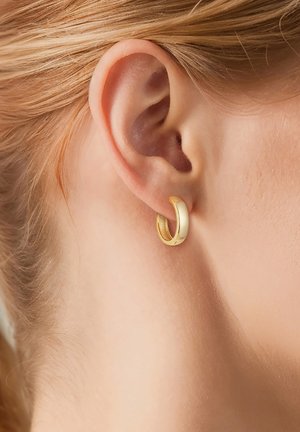 Primer plano de la oreja de una persona rubia que lleva un pequeño y liso pendiente de aro dorado brillante.