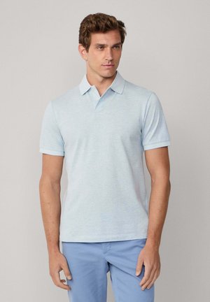 Uomo che indossa una polo azzurro chiaro e pantaloni blu, in piedi di fronte a uno sfondo grigio uniforme, che guarda lievemente di lato.