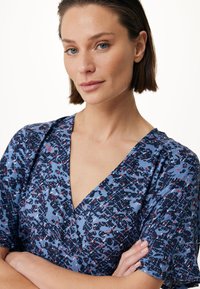 Květinové šaty s úzkým páskem v modré barvě s abstraktními vzory v námořnické modři, červené a bílé. Mají hluboký výstřih do V a krátké, mírně nabrané rukávy.