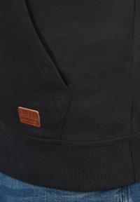 Sudadera negra con capucha, fabricada en un tejido texturizado, con bajo acanalado y una etiqueta de cuero marrón con la palabra "BLEND" cosida en ella.