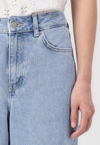 Ljust blå denimjeans med hög midja, med knappstängning, sidofickor och subtila bleknade partier längs tygets struktur.