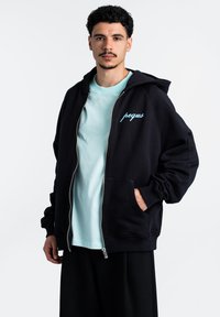 Juodas zip-up džemperis su kengūros kišene, išsiskiriantis šviesiai mėlynu siuvinėtu tekstu. Nešiojamas virš šviesiai mėlynos marškinėlių, rodoma neutralioje fone.