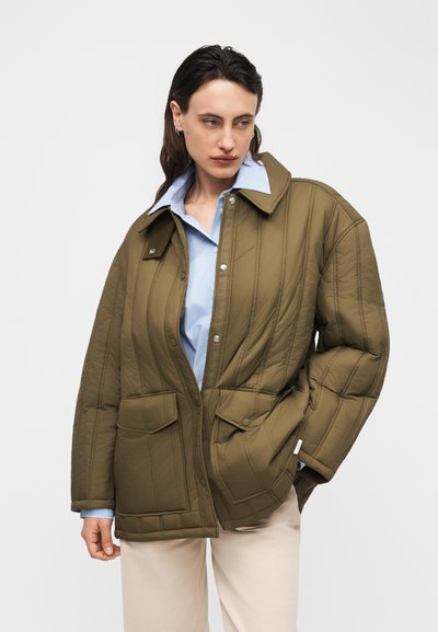 Γυναίκα που φοράει υπερμεγέθη καπιτονέ σακάκι σε ελιά, πάνω από ανοιχτό μπλε πουκάμισο με γιακά και μπεζ παντελόνι, στέκεται μπροστά σε λευκό φόντο.