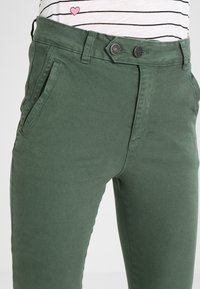 Zielone bawełniane spodnie o dopasowanym kroju, z dwiema kieszeniami z przodu i detalem w postaci podwójnego zapięcia w pasie, o gładkiej fakturze i bez widocznych wzorów.