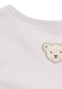 Helles rosa T-Shirt mit kurzen Ärmeln und rundem Ausschnitt, mit einem beige gestickten Bären-Patch auf der linken Brust. Glatte Stoffstruktur.