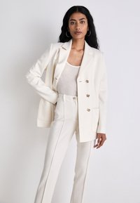 Cremfarbener maßgeschneiderter Blazer mit doppelt-knöpfbaren Knöpfen, kombiniert mit schmal geschnittenen Hosen. Getragen über einem hellgrauen Oberteil mit glatter Textur.